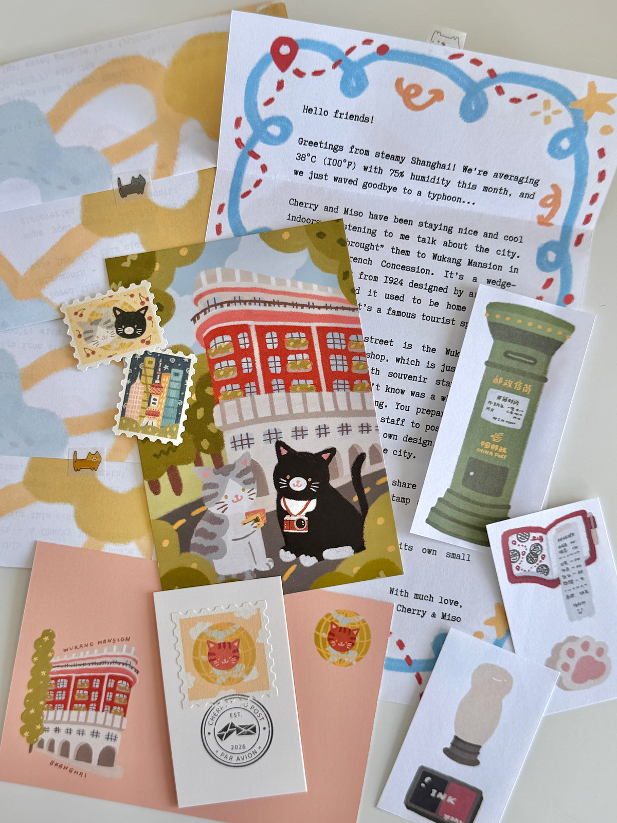 A flatlay on a white table of cat and post office-themed stationery items, including a letter, postcard, stamp stickers, postmark card, paper scraps, and an envelope.