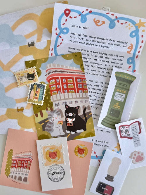 A flatlay on a white table of cat and post office-themed stationery items, including a letter, postcard, stamp stickers, postmark card, paper scraps, and an envelope.