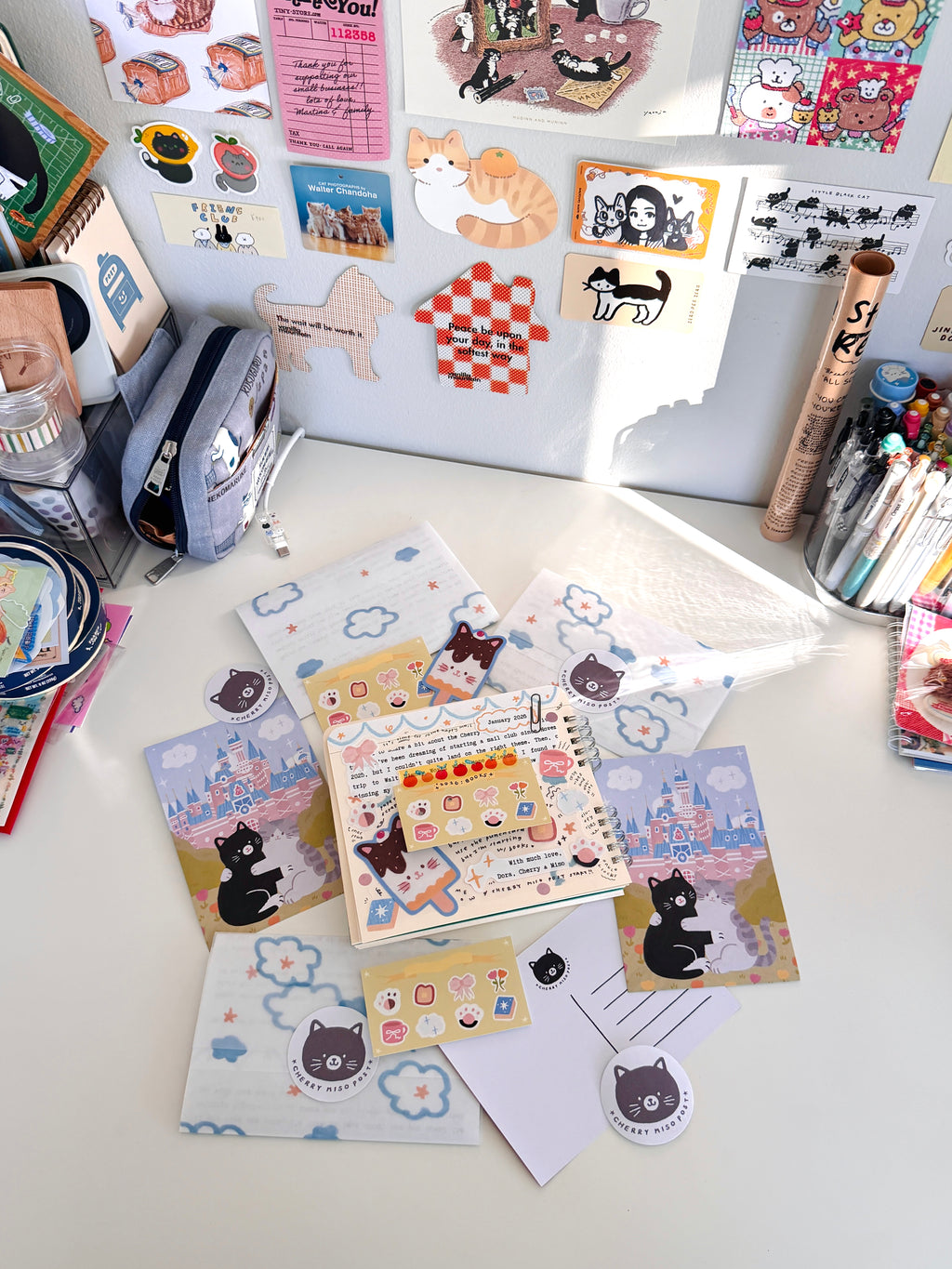 Flat lay of colorful stationery including postcards, stickers, and paper with cute cat and cloud designs from the January edition of Cherry Miso Post displayed on a white desk with a wall decorated in various small art prints and stickers.