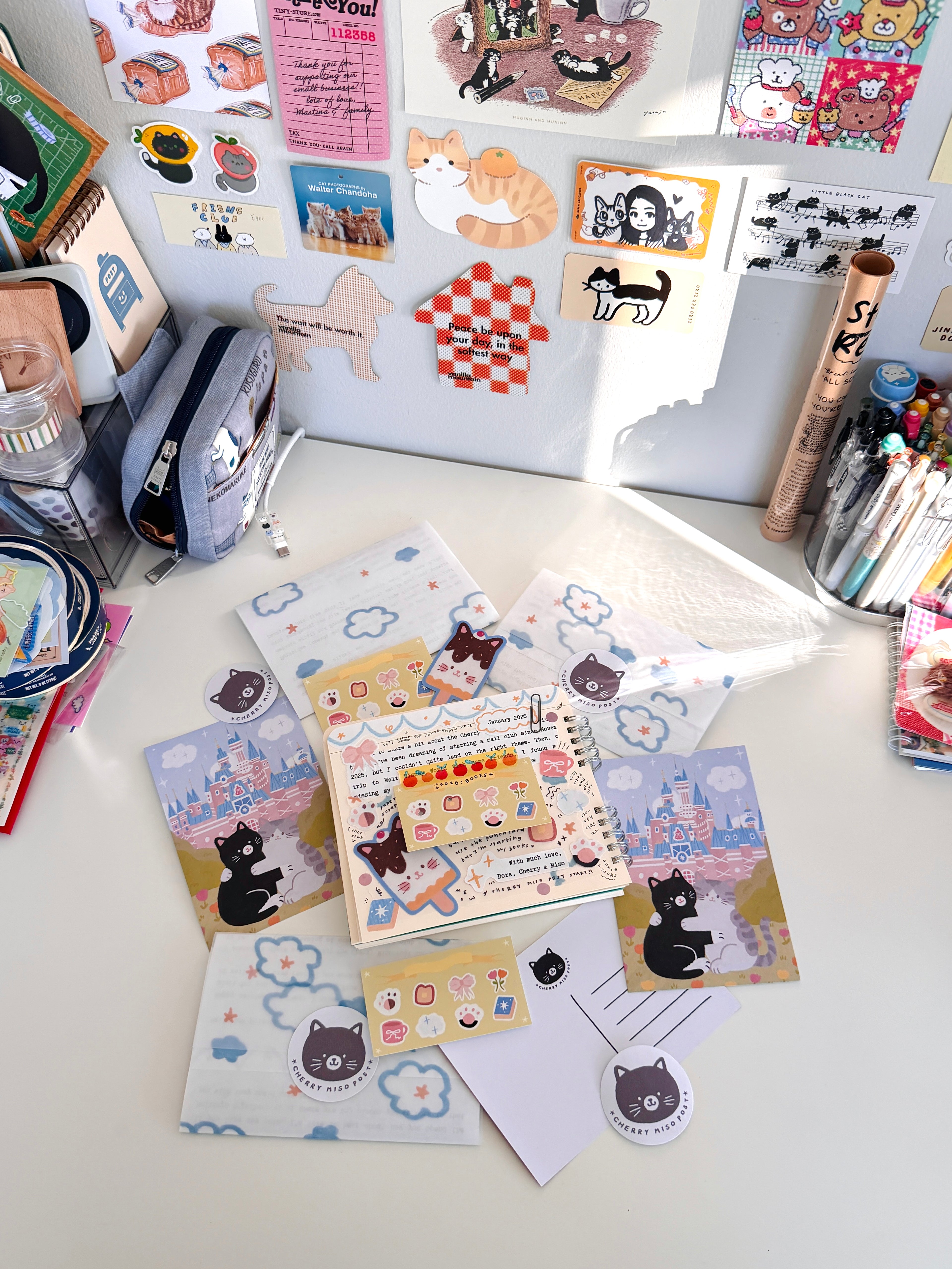 Flat lay of colorful stationery including postcards, stickers, and paper with cute cat and cloud designs from the January edition of Cherry Miso Post displayed on a white desk with a wall decorated in various small art prints and stickers.