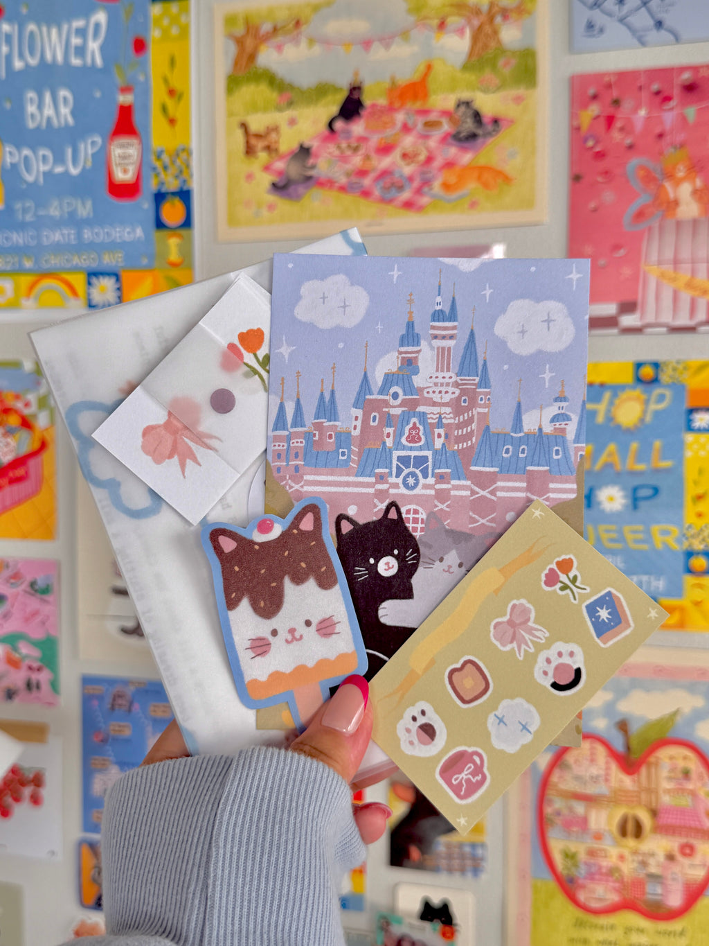Hand holding a collection of colorful stationery including a postcard with a whimsical castle, cat-themed stickers, and cute paper items against a vibrant wall of postcards.