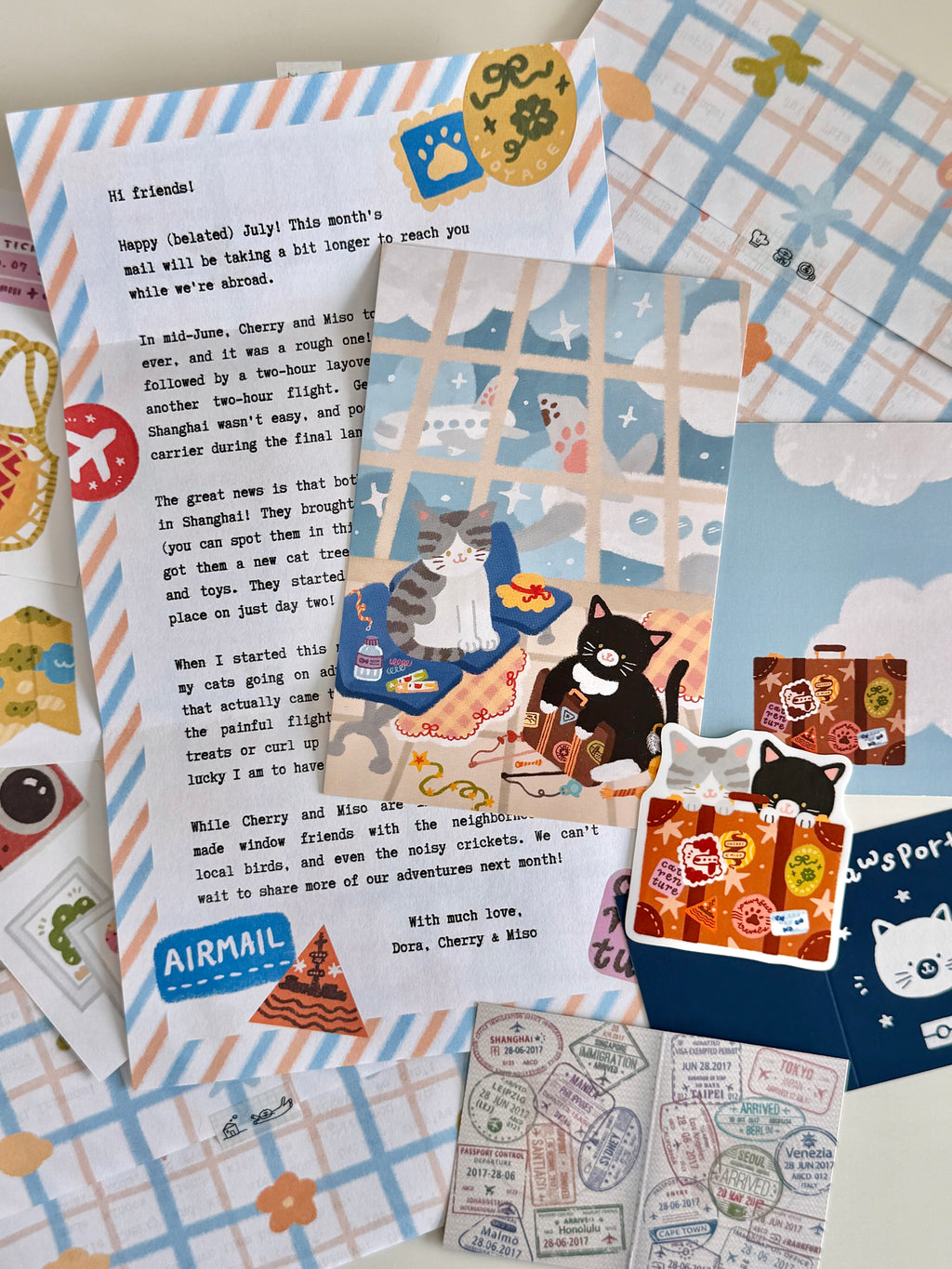 A flatlay on a white table of cat and travel-themed stationery items, including a letter, postcard, sticker, paper passport, and paper scraps with illustrations.