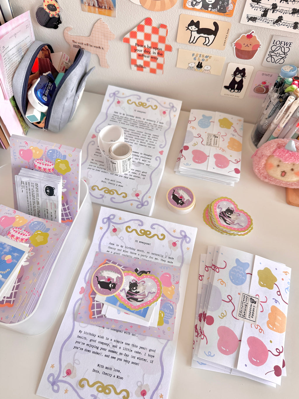 Stacks of monthly letters being folded with postcards, stickers, polaroid cards, and paper scraps wrapped inside.