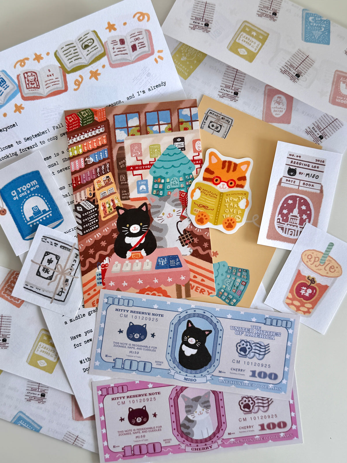 A flatlay on a white table of cat and book-themed stationery items, including a letter, postcard, sticker, paper money, paper scraps, and an envelope.